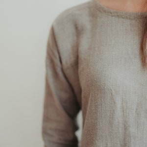 May include: A close-up of a light brown linen shirt with a round neckline. The fabric has a textured, woven appearance.