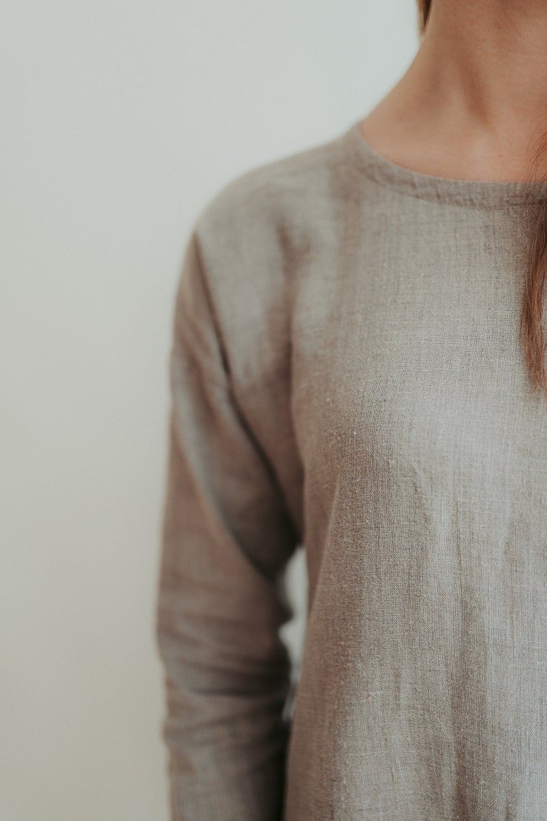 May include: A close-up of a light brown linen shirt with a round neckline. The fabric has a textured, woven appearance.