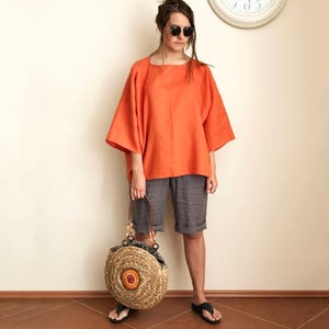 K&ouml;nnte beinhalten: Eine Frau tr&auml;gt ein locker sitzendes, orangefarbenes Leinenoberteil und graue Leinenhosen. Sie tr&auml;gt eine runde, geflochtene Strohtasche mit bunten Akzenten.