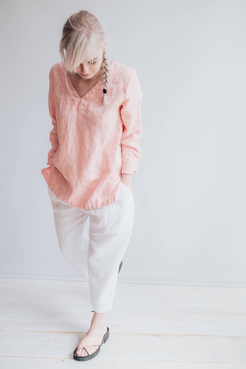 May include: A woman wearing a light pink linen top with long sleeves and white linen pants. She is wearing sandals and has her blonde hair in a braid.
