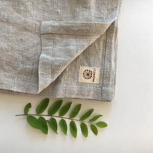 May include: A gray linen kitchen towel with a brown tag that says "Linenbee" with a stylized flower design. The towel is folded in half and a green leafy branch is in the bottom right corner of the image.