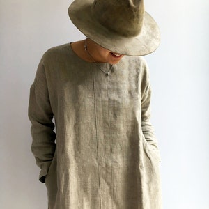 A woman is wearing a linen tunic made from natural taupe color linen. The tunic has long sleeves and the woman has put her hands in the pockets. Pockets are placed in the side seams. The tunic has a round neckline and runs past the hips.