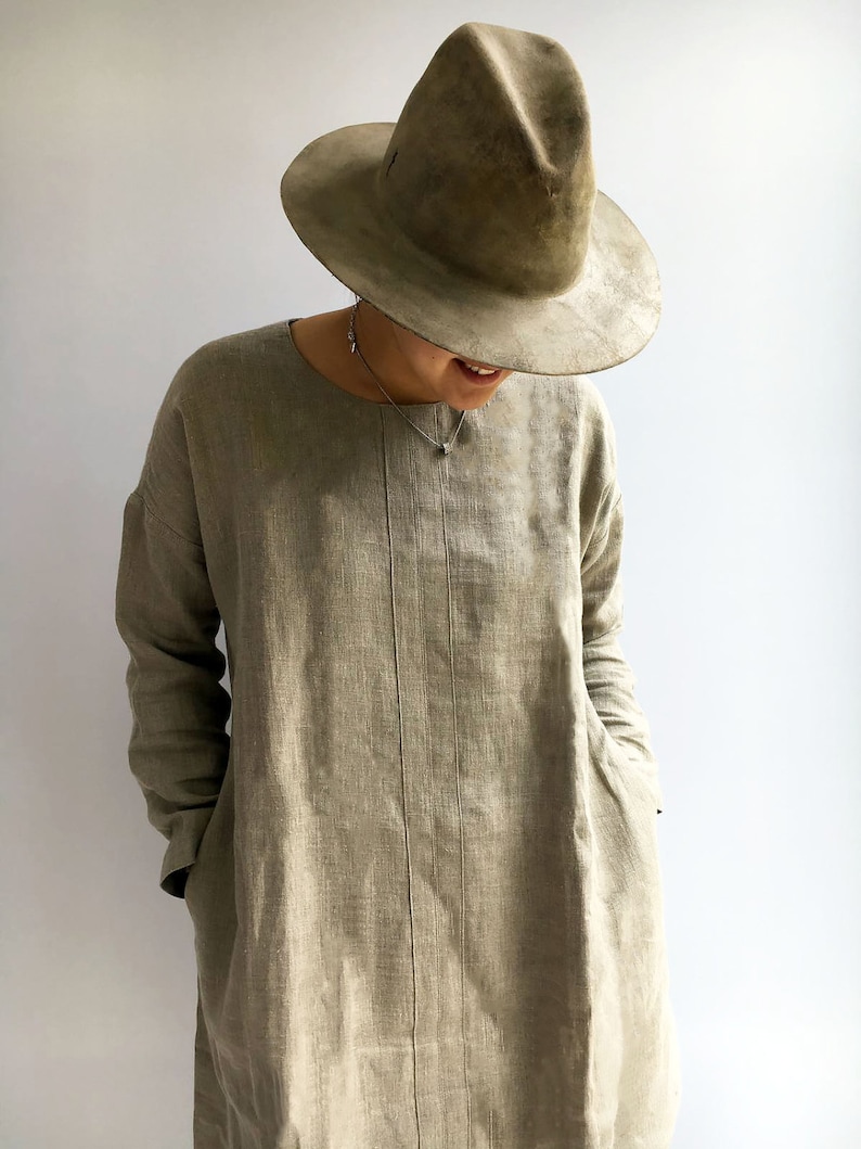 A woman is wearing a linen tunic made from natural taupe color linen. The tunic has long sleeves and the woman has put her hands in the pockets. Pockets are placed in the side seams. The tunic has a round neckline and runs past the hips.