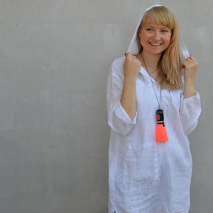 Op de afbeelding: Een vrouw draagt een witte linnen tuniek met een capuchon en een felroze kwastketting. Ze draagt lichtblauwe jeans en heeft haar capuchon opgetrokken.