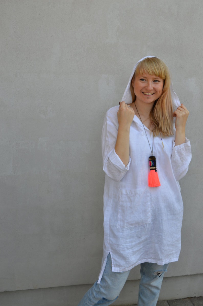 Op de afbeelding: Een vrouw draagt een witte linnen tuniek met een capuchon en een felroze kwastketting. Ze draagt lichtblauwe jeans en heeft haar capuchon opgetrokken.