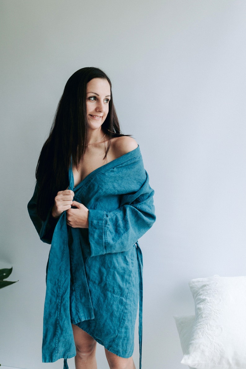 May include: A woman wearing a teal blue linen robe with a long sash. The robe is open in the front, revealing her shoulders and chest. She is standing in front of a white wall.
