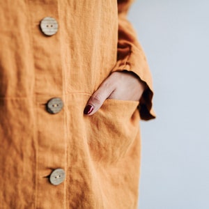 May include: A close-up of a person wearing a long, orange linen jacket with three wooden buttons. The jacket has a pocket on the left side and the person's hand is in the pocket.