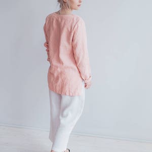 May include: A woman wearing a pink linen top and white linen pants. She is standing in front of a white wall and looking to the right.