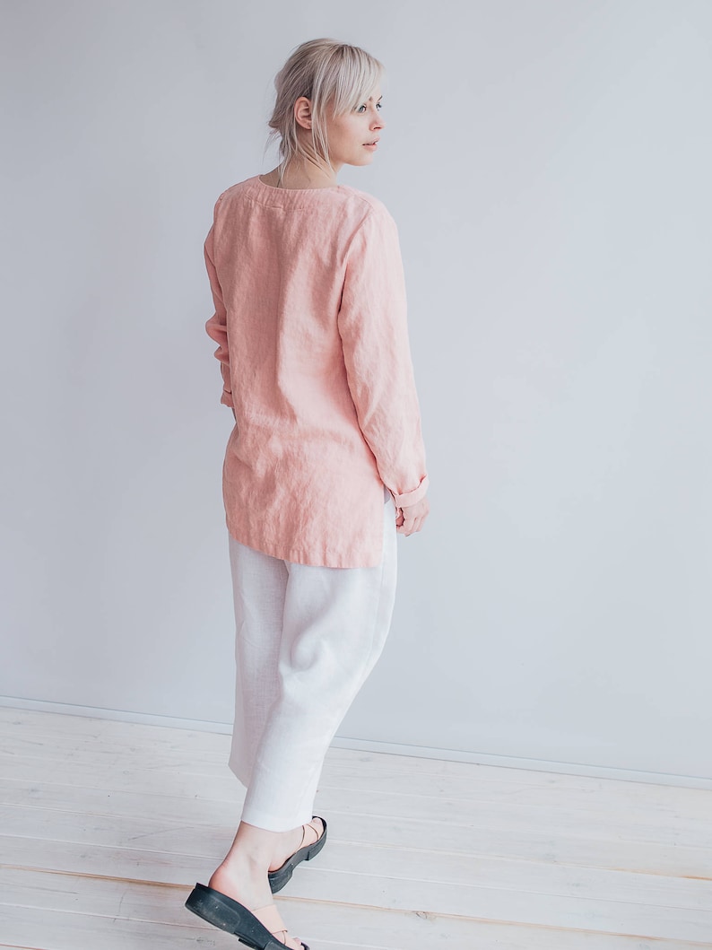 May include: A woman wearing a pink linen top and white linen pants. She is standing in front of a white wall and looking to the right.