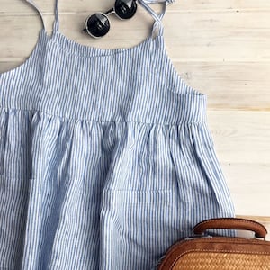 May include: A blue and white striped linen dress with thin straps and two pockets. The dress is laying on a white wooden surface with a brown woven bag and round sunglasses.