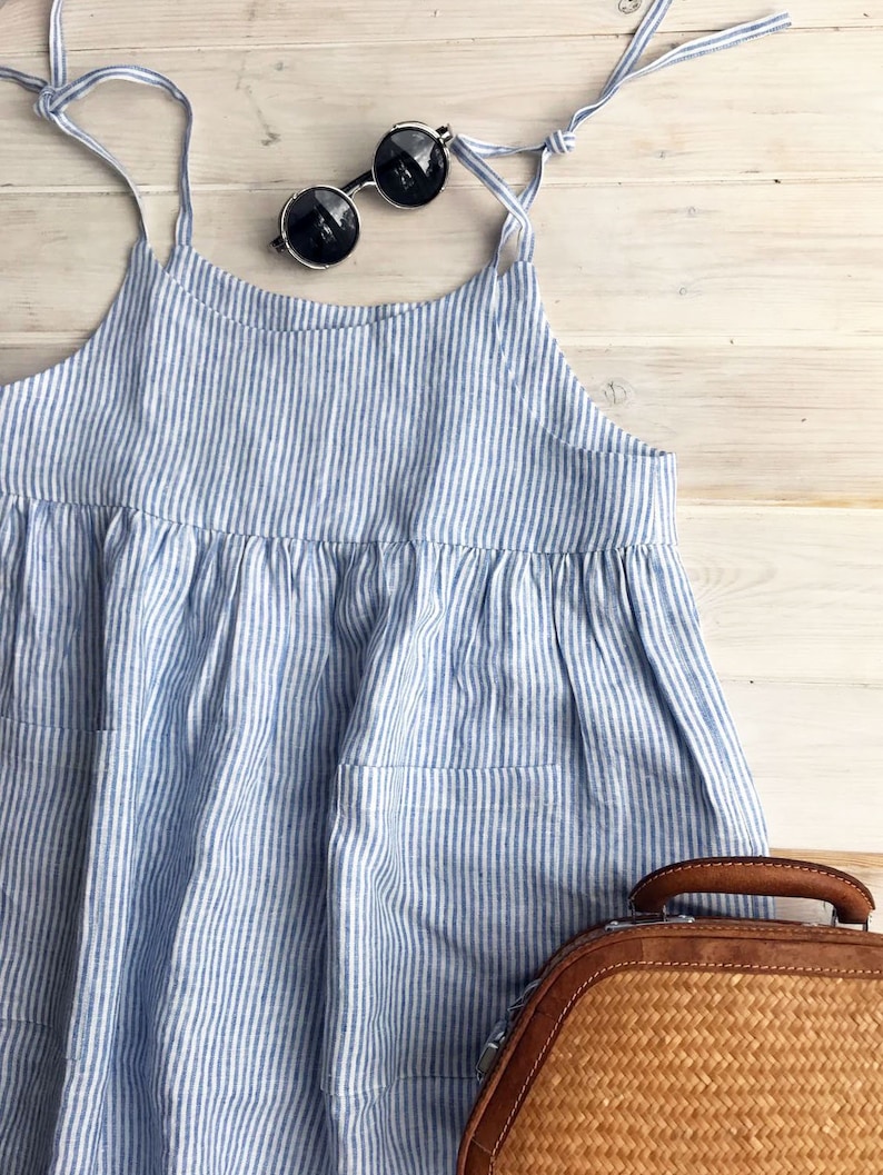 May include: A blue and white striped linen dress with thin straps and two pockets. The dress is laying on a white wooden surface with a brown woven bag and round sunglasses.