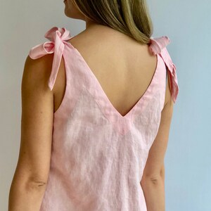 May include: A light pink linen top with a V-neck and tie shoulder straps. The straps are tied into bows. The top is sleeveless and appears to be made of a lightweight fabric.