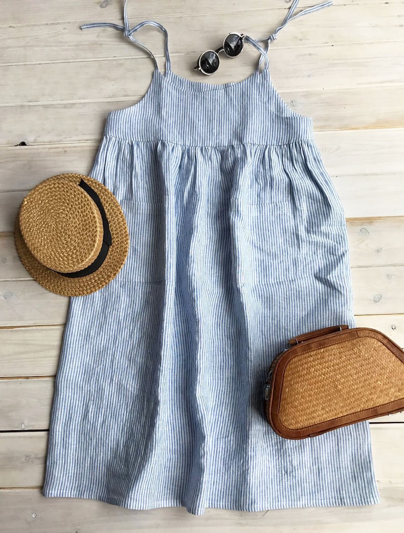 May include: A blue and white striped linen dress with thin straps. The dress has a loose fit and a gathered waist. It is photographed on a white wooden background with a straw hat and a woven bag.