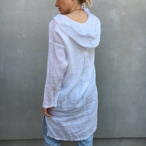 Op de afbeelding: Een witte linnen tuniek met een capuchon. De tuniek heeft lange mouwen en een losse pasvorm. Het model draagt de tuniek met een blauwe jeans.