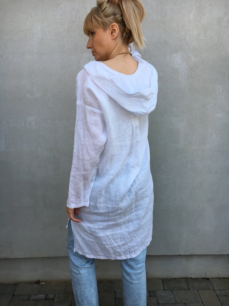 Op de afbeelding: Een witte linnen tuniek met een capuchon. De tuniek heeft lange mouwen en een losse pasvorm. Het model draagt de tuniek met een blauwe jeans.