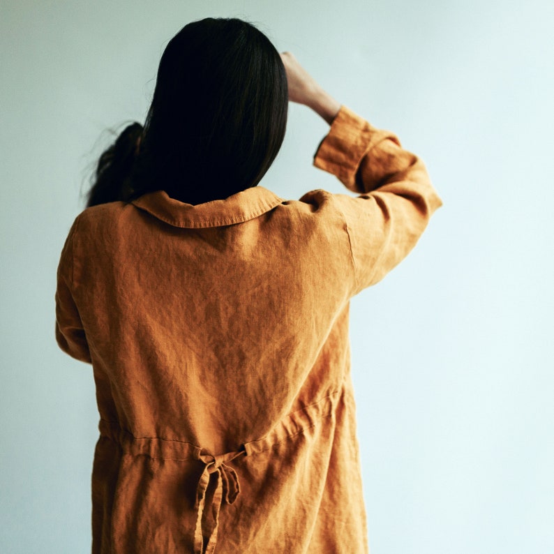 May include: A woman wearing a long, mustard yellow linen shirt with a drawstring waist. The shirt has a relaxed fit and a collar.