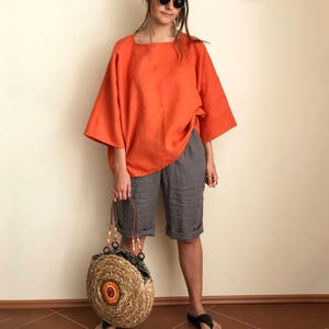 K&ouml;nnte beinhalten: Eine Frau tr&auml;gt ein locker sitzendes orangefarbenes Leinenoberteil, graue Leinenhosen und schwarze Sandalen. Sie tr&auml;gt eine runde, geflochtene Strohtasche mit orangefarbenen und braunen Akzenten.