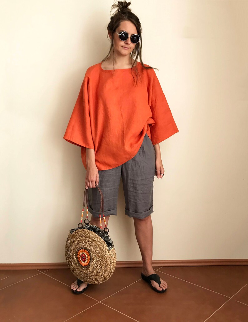 K&ouml;nnte beinhalten: Eine Frau tr&auml;gt ein locker sitzendes orangefarbenes Leinenoberteil, graue Leinenhosen und schwarze Sandalen. Sie tr&auml;gt eine runde, geflochtene Strohtasche mit orangefarbenen und braunen Akzenten.