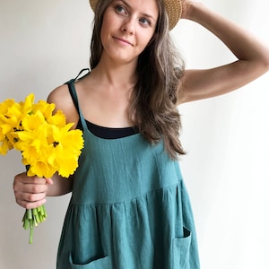 May include: A woman wearing a teal linen dress with pockets and a straw hat holds a bouquet of yellow daffodils.