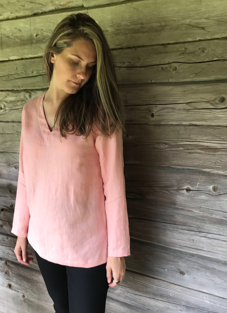 May include: A pale pink linen blouse with a v-neck and long sleeves. The blouse is worn by a person standing in front of a wooden wall.