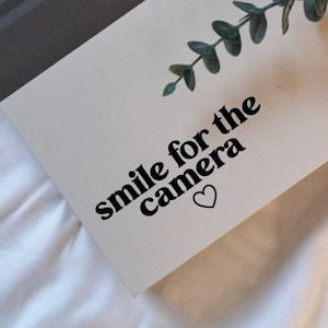 May include: White notebook with a black and white decal that says "smile for the camera" with a small heart. The notebook is on a white surface with a green plant.