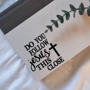May include: White background with a grey and white notebook. The notebook has a black vinyl decal that says "Do you follow Jesus this close" with a black cross.