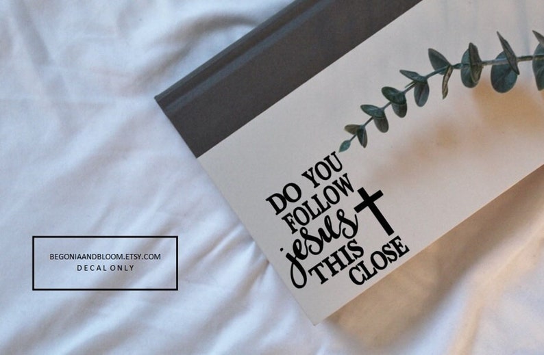 May include: White background with a grey and white notebook. The notebook has a black vinyl decal that says "Do you follow Jesus this close" with a black cross.