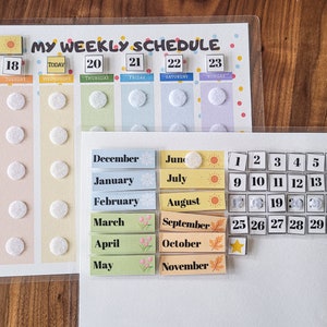 May include: Two clear plastic dry-erase boards with a weekly schedule and a calendar. The weekly schedule board has a colourful background with days of the week and dates. The calendar board has a white background with numbers 1 through 31. Both boards have removable stickers with months of the year and icons.