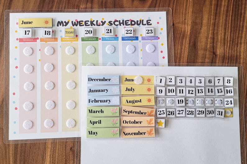 May include: Two clear plastic dry-erase boards with a weekly schedule and a calendar. The weekly schedule board has a colourful background with days of the week and dates. The calendar board has a white background with numbers 1 through 31. Both boards have removable stickers with months of the year and icons.