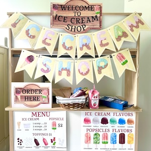 Può includere: Un'esposizione di gelateria in legno con uno striscione che recita "ICE CREAM SHOP". Il negozio comprende un menu, gusti di gelato, gusti di ghiaccioli e un cartello "ORDER HERE". Il negozio ha un cestino di coni e una bottiglia di sciroppo di fragole.