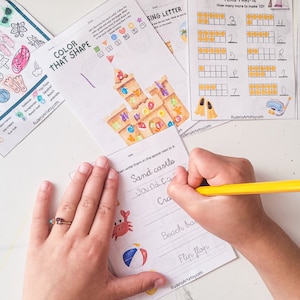 May include: A child's hand writes the words "Sand castles" on a worksheet with a picture of a beach ball and a crab. The worksheet is part of a set of summer-themed educational activities.