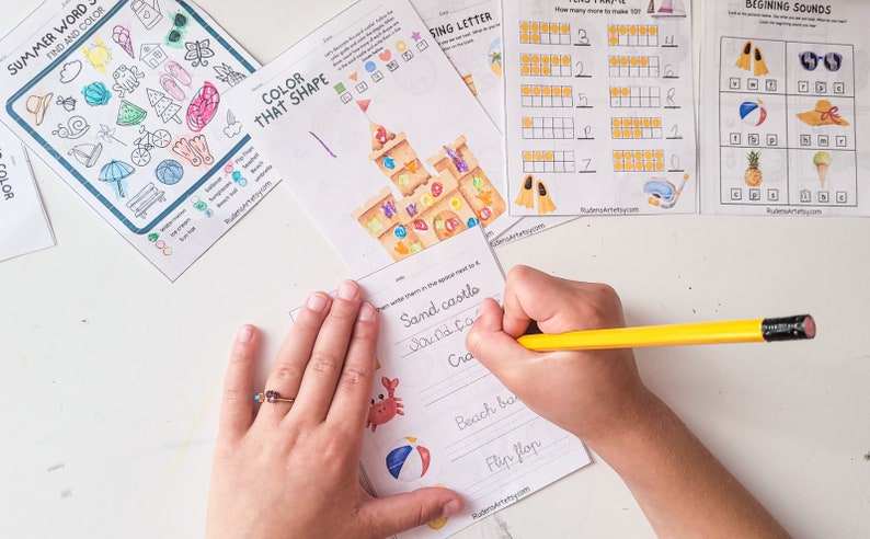 May include: A child's hand writes the words "Sand castles" on a worksheet with a picture of a beach ball and a crab. The worksheet is part of a set of summer-themed educational activities.