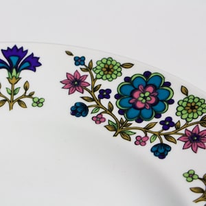May include: A white ceramic plate with a floral design. The design features blue, green, pink, and gold flowers and leaves.