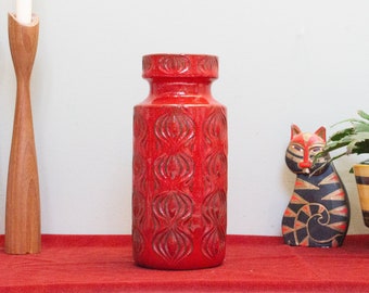 May include: A red ceramic vase with a geometric pattern. The vase is tall and cylindrical with a slightly flared top.