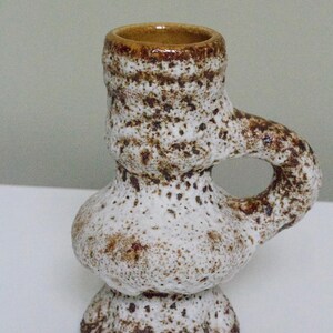 May include: A white ceramic candle holder with brown speckles and a handle. The holder has a rounded base and a wide opening at the top.