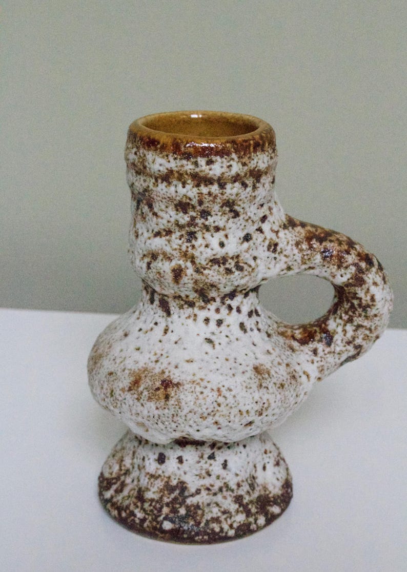 May include: A white ceramic candle holder with brown speckles and a handle. The holder has a rounded base and a wide opening at the top.