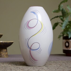 May include: White ceramic vase with a stylized abstract design in blue, yellow, and pink. The vase is oval shaped and has a smooth, glossy finish.