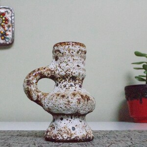 May include: A white ceramic vase with brown speckles and a unique handle. The vase has a textured surface and a rounded base.