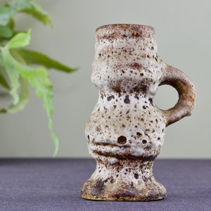 May include: A small, white ceramic vase with a brown glaze and a textured surface. The vase has a rounded body and a single handle.