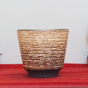 May include: A brown and black ceramic planter with a textured, striped design. The planter has a wide, round opening and a slightly flared base.