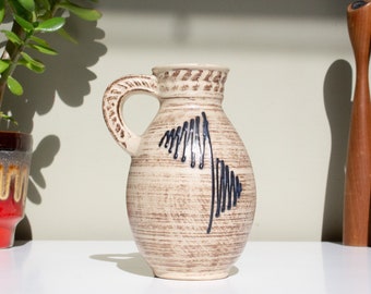 May include: A cream-coloured ceramic jug with a brown glaze and a blue and black abstract design. The jug has a handle and a rounded body.