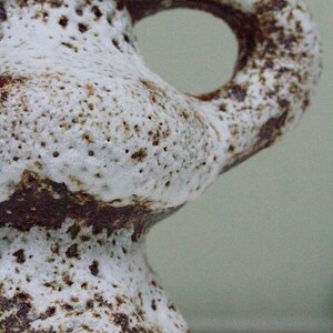 May include: A white ceramic vase with brown speckles and a unique, organic shape. The vase has a wide base and a narrow neck with a handle.