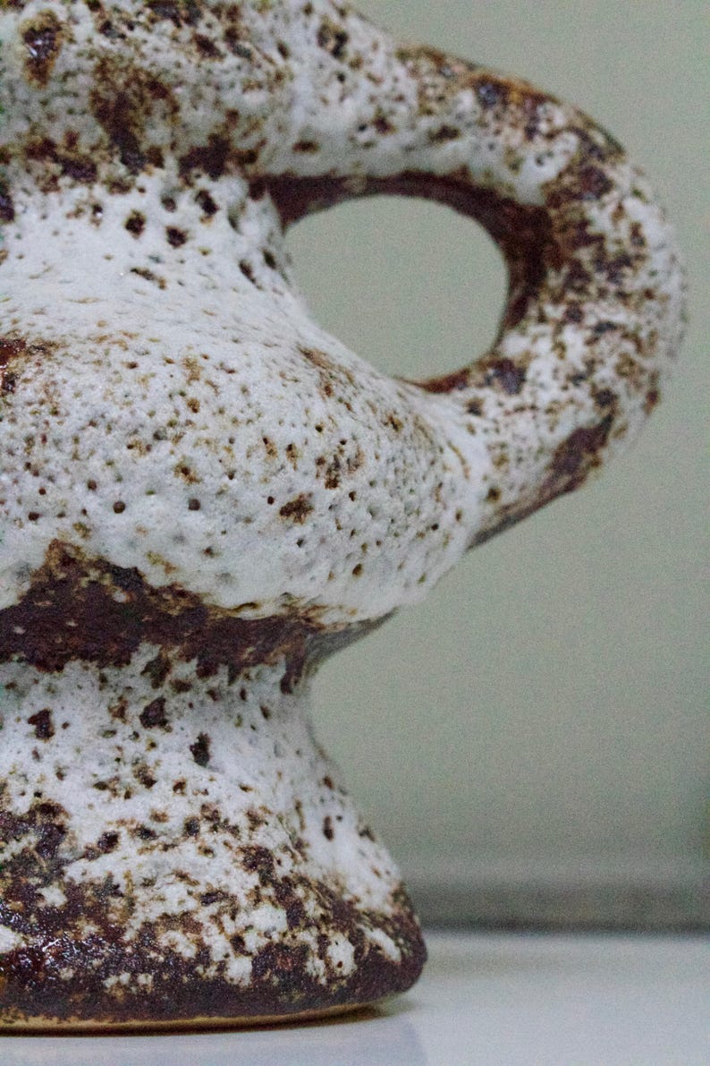 May include: A white ceramic vase with brown speckles and a unique, organic shape. The vase has a wide base and a narrow neck with a handle.