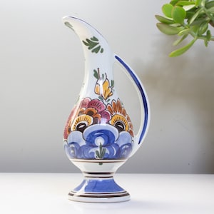 May include: A white ceramic pitcher with a blue, orange, and yellow floral design. The pitcher has a wide base and a curved spout.