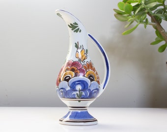 May include: A white ceramic jug with a blue, orange and yellow floral design. The jug has a wide base and a curved spout.