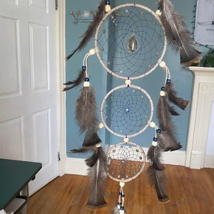 May include: A triple dream catcher with beige and blue string, brown feathers, and wooden beads. The top dream catcher has a blue stone pendant.