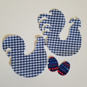 Op de afbeelding: Drie stoffen uitsparingen: twee hanenvormen in blauw-wit vichyruit en twee patronen eivormen met rode en blauwe stippen. De vormen zijn gerangschikt op een witte achtergrond, wat wijst op een knutsel- of Paasthema.