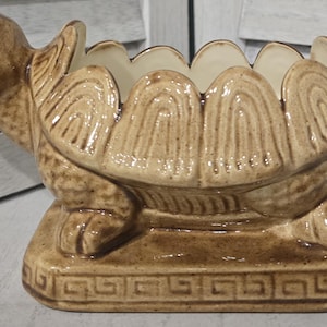 Könnte beinhalten: Ein schildkrötenförmiger Übertopf aus Keramik mit einer glänzenden, braun und beige gesprenkelten Glasur. Der Panzer hat einen gewellten Rand und der rechteckige Sockel ist mit einem geometrischen Mäander-Muster verziert. Das Innere ist cremefarben.