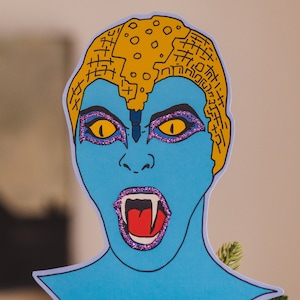 May include: A blue paper cutout of a monster with a yellow and black patterned head, open mouth with fangs, and purple glitter around the eyes.