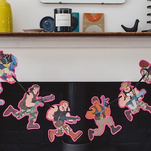 May include: A pink and black paper garland with characters from the movie Predator. The characters are running and holding weapons. The garland is hanging from a fireplace mantel.